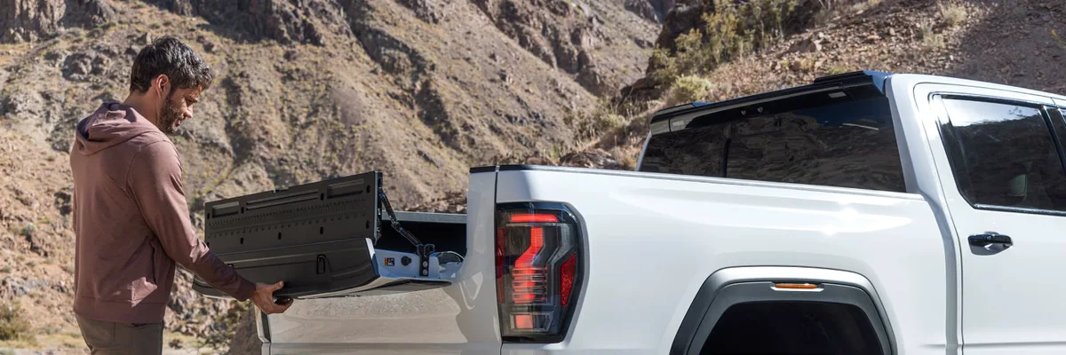 A Man Closing the Multi-Function Tailgate of a White GMC Sierra 1500 AT4X Truck on a Dirt Road in a Mountainous Landscape